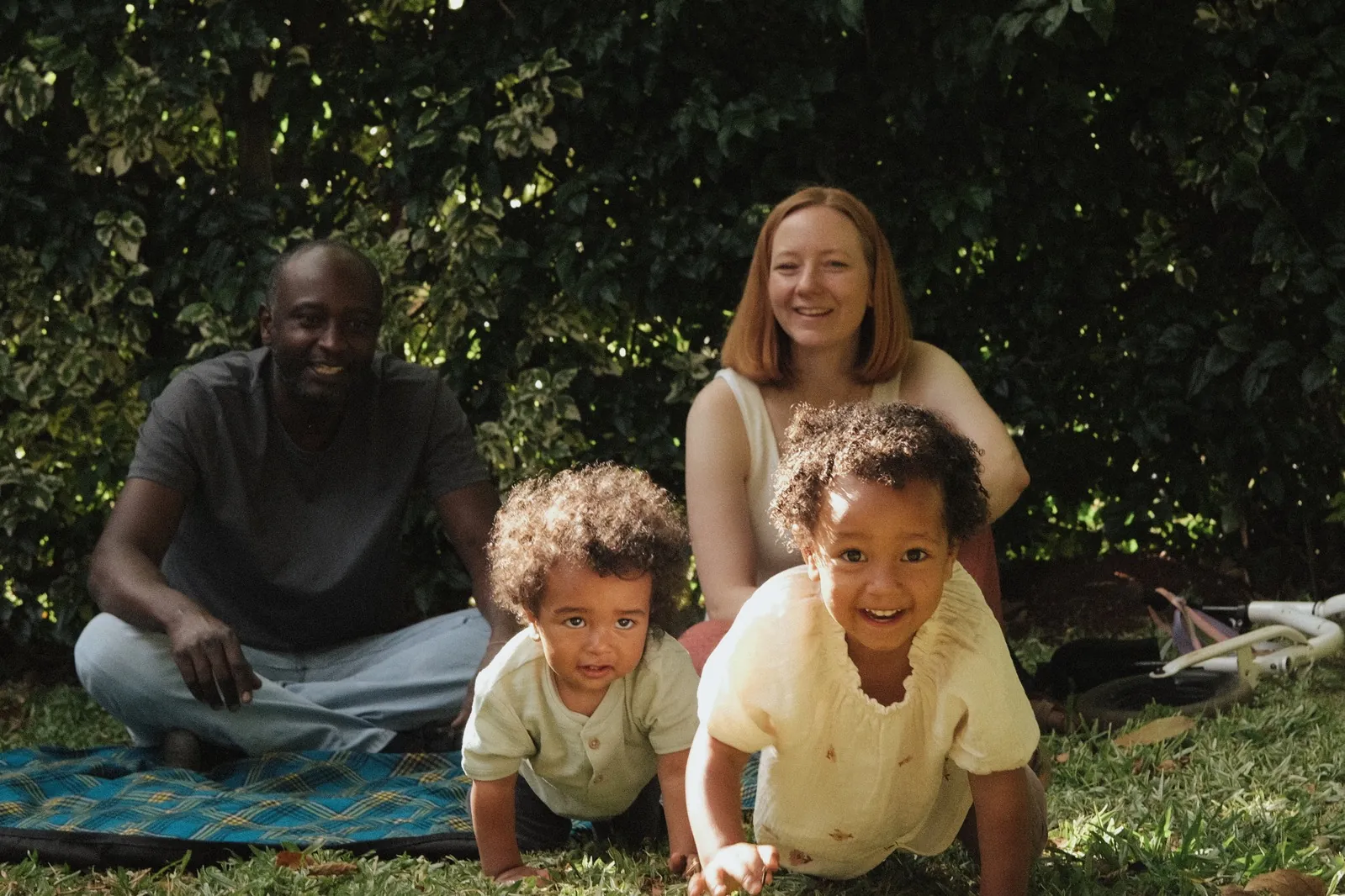 Two young children crawling towards the camera while their parents laugh behind them in a Nairobi garden