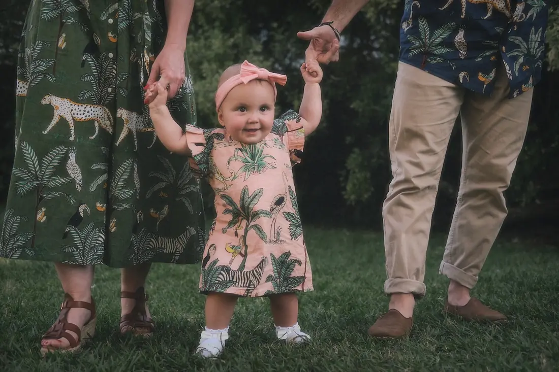 A young child taking first steps while holding two adults' hands